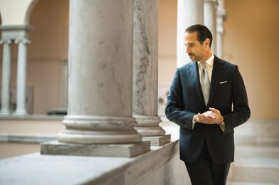 Brian Sacawa wearing a tailored navy suit in a marble-columned interior for Michael Andrews Bespoke’s 20 for 20 Look Book.