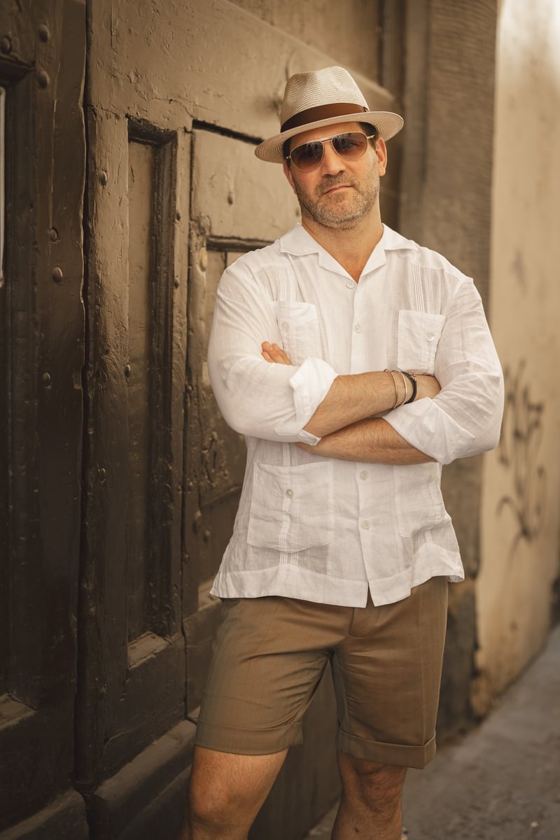 Michael wearing a white double-pocket linen camp shirt and tailored Solaro shorts, styled with a straw fedora and sunglasses